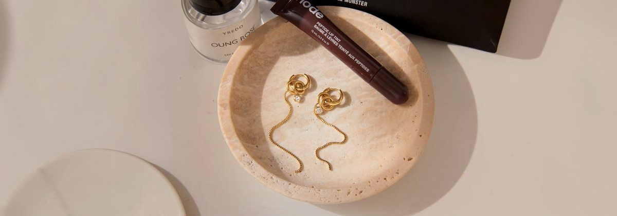 A pair of gold dangle earrings with coiled, snake-like drops rests in a round beige travertine stone dish. A dark brown makeup stick and a small bottle labeled 'COUTURE' are placed nearby on a white surface, with a black box in the background.