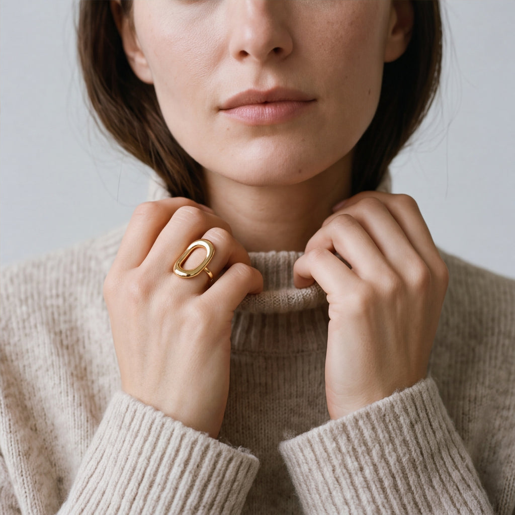 A close-up lifestyle shot of a model wearing a amethyst & sable axis frame ring in gold with a large open oval design on her finger, wearing a beige knit turtleneck sweater against a soft gray background.