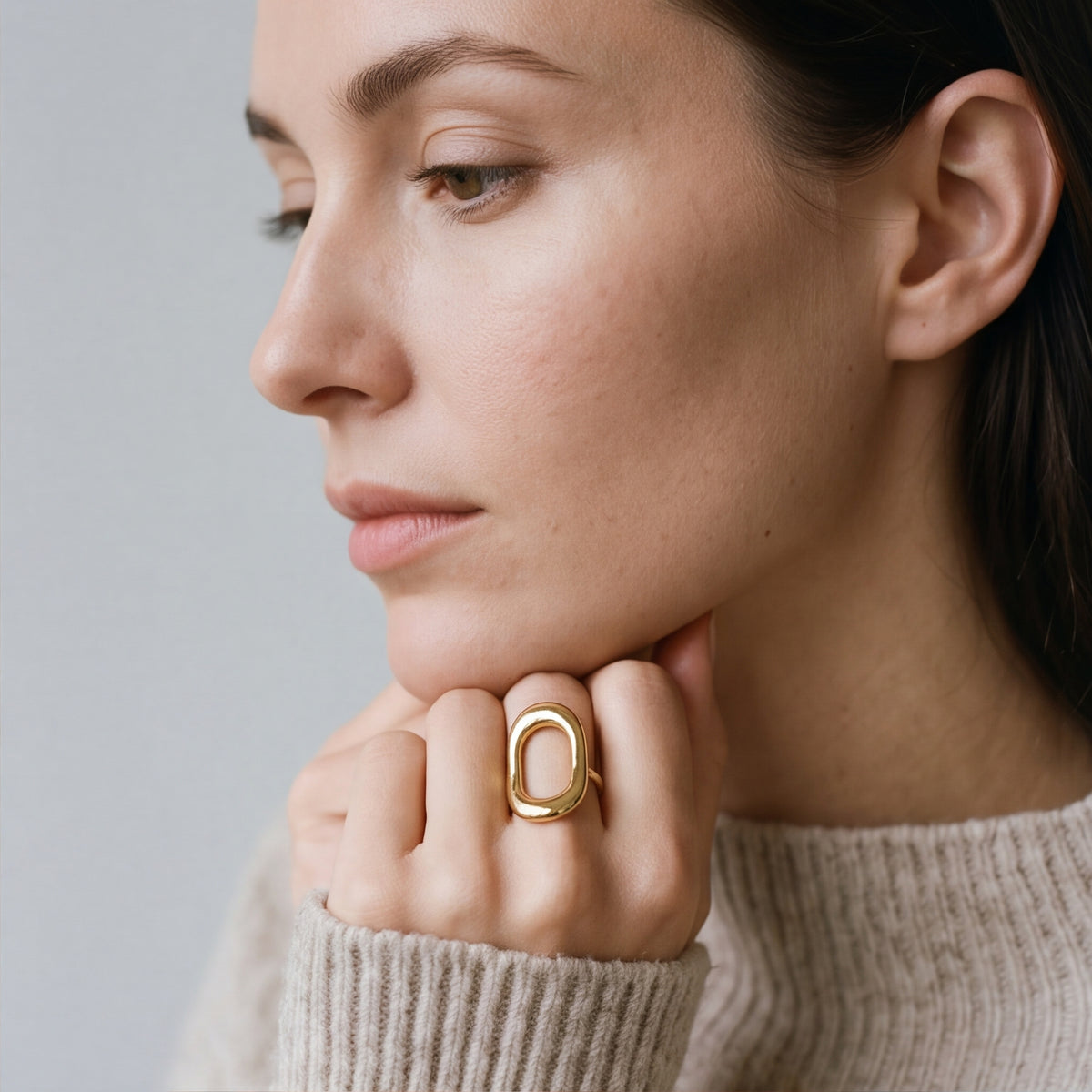 A close-up lifestyle shot of a model wearing a amethyst & sable axis frame ring in gold with a large open oval design on her finger, wearing a beige knit turtleneck sweater against a soft gray background.