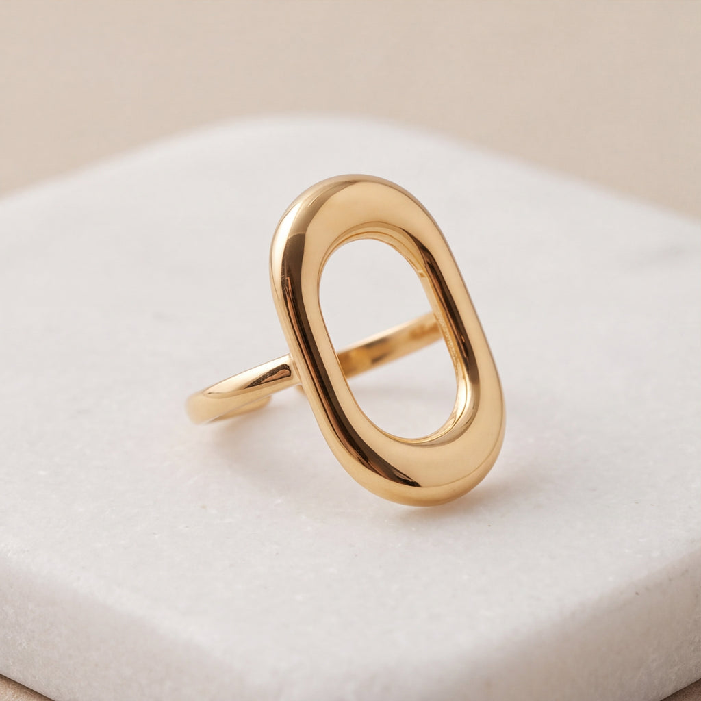 A polished amethyst & sable axis frame ring in gold with a large, open oval design, displayed on a textured white square pedestal against a soft beige background.