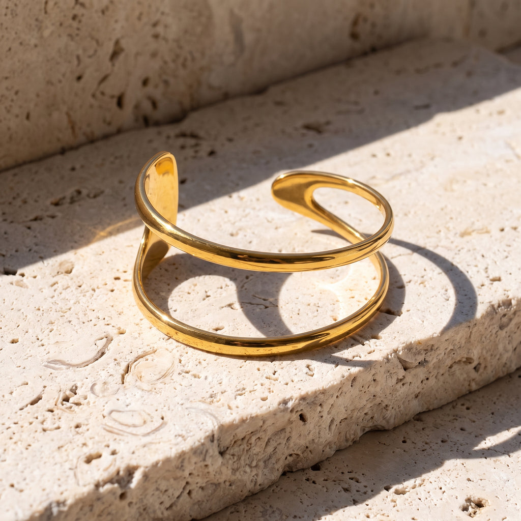 A polished gold axis strata open cuff bracelet with a smooth, curved design, resting on a textured beige stone surface under bright natural sunlight.