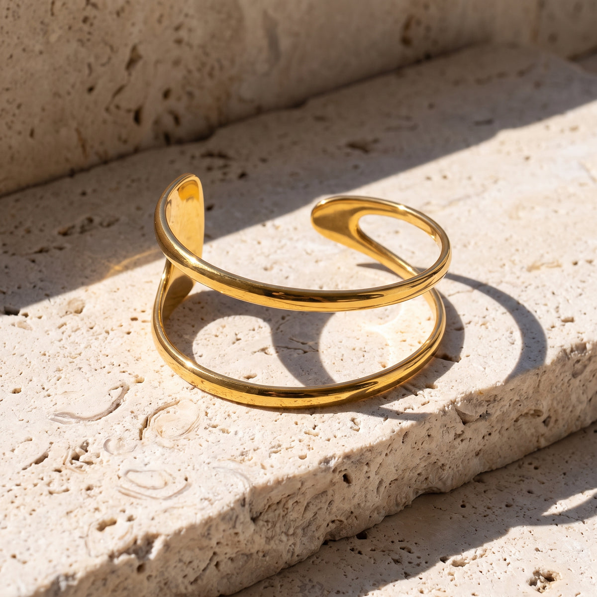 A polished gold axis strata open cuff bracelet with a smooth, curved design, resting on a textured beige stone surface under bright natural sunlight.