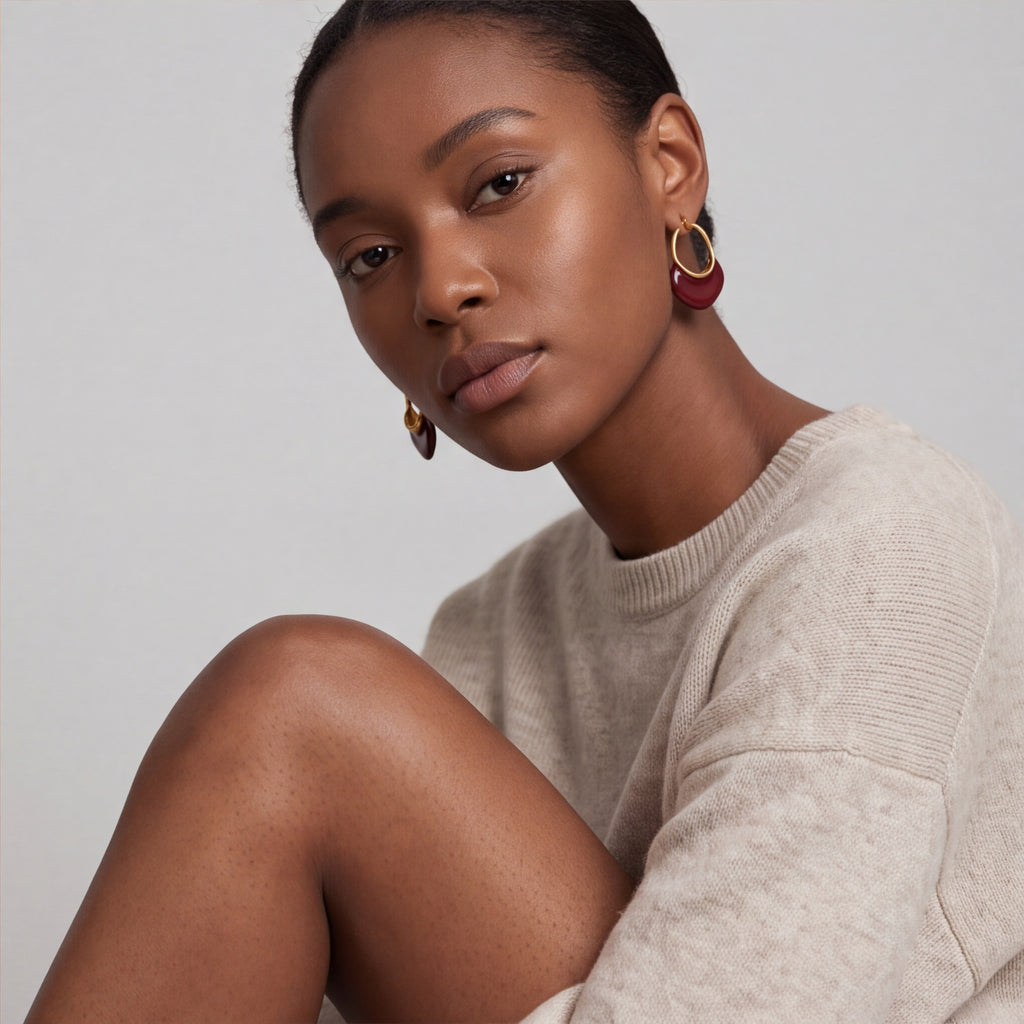 A close-up of a model wearing calypso camille hoop earrings with deep red circular inserts, paired with a light beige knit sweater against a neutral gray background.