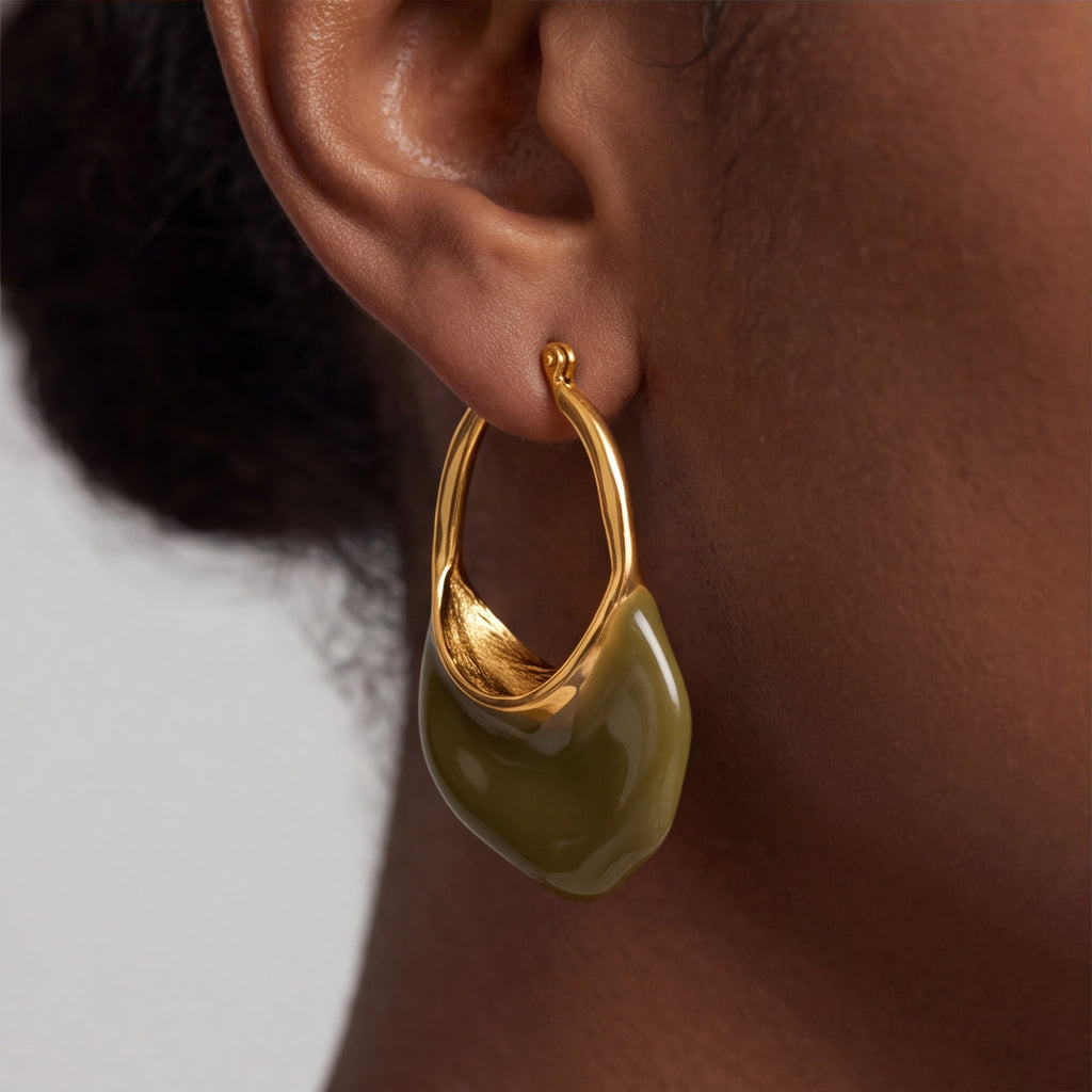 A close-up of a person's ear wearing calypso camille abstract gold hoop earring with a glossy olive green drop, shown against a neutral light gray background.
