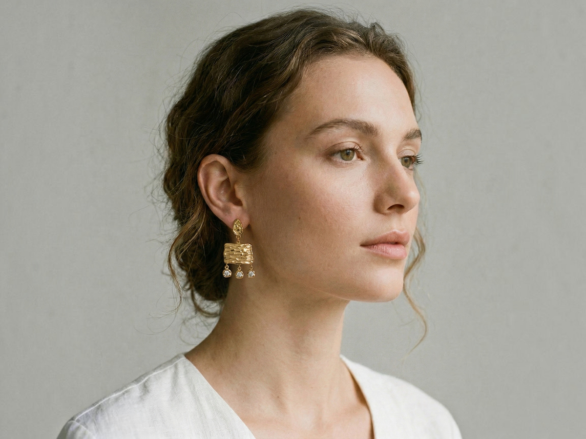 A woman in a white top wearing a gold textured geometric dangle earring with small pearl-like drops, shown in a close-up profile against a neutral gray background.