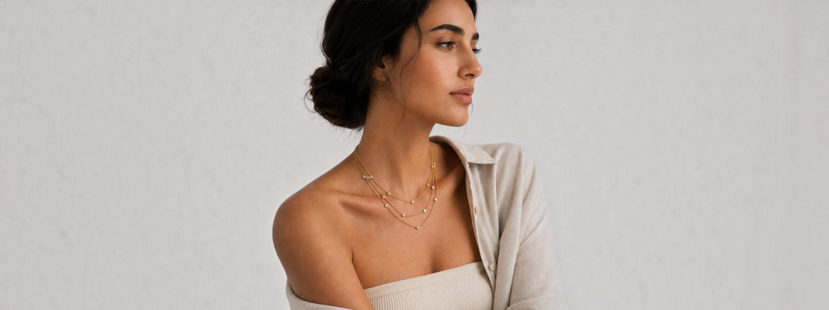 A woman with her hair in a low bun is shown in profile wearing a delicate layered gold necklace with small bead details. She is wearing a light beige off-the-shoulder top and a light beige overshirt against a neutral, textured off-white background.